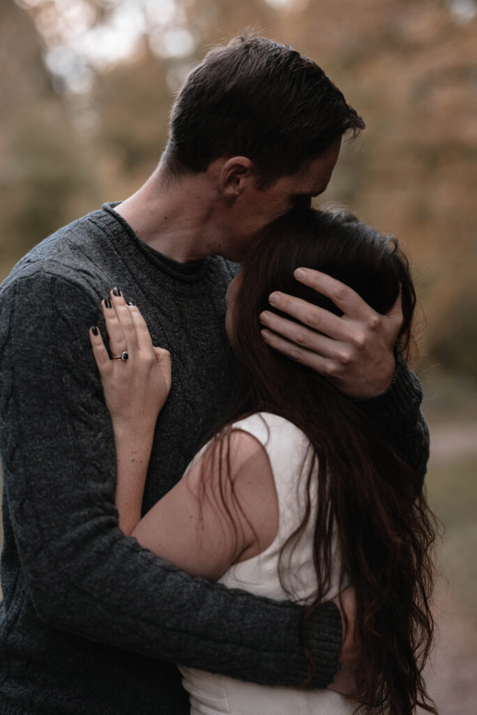 Man holding woman close to him, his hand in her hair. Her hand with engagement ring is placed onto his arm.