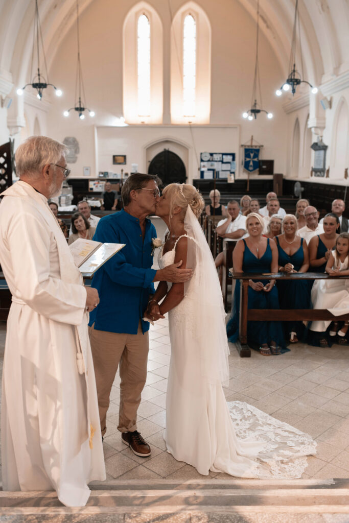 Bride and Groom having their first kiss at their wedding ceremony surrounded by smiling friends and family