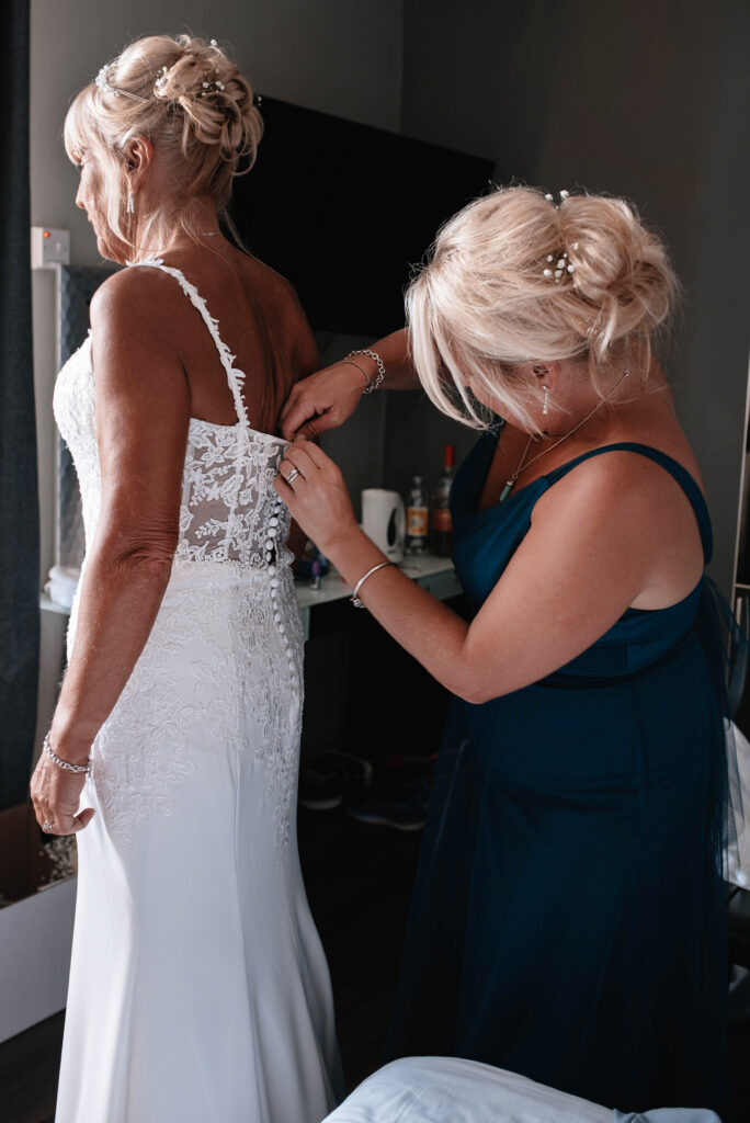 Maid of honor dressed in long blue dress fastening buttons on the brides dress