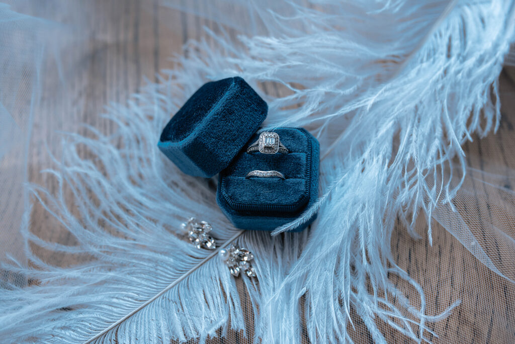 An engagement ring and wedding band sit in a velvet ring box with earrings and a feather in the background on top of a wooden table