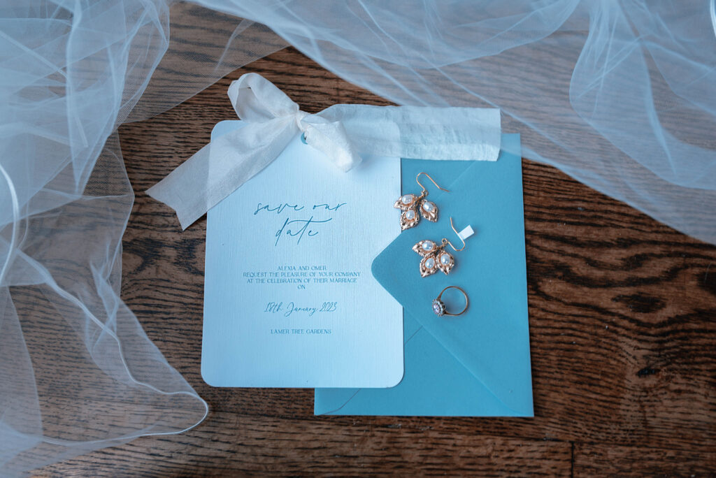 A save the date card and envelope are laid on a wooden table, on top is a pair of earrings and an engagement ring, around them is a veil