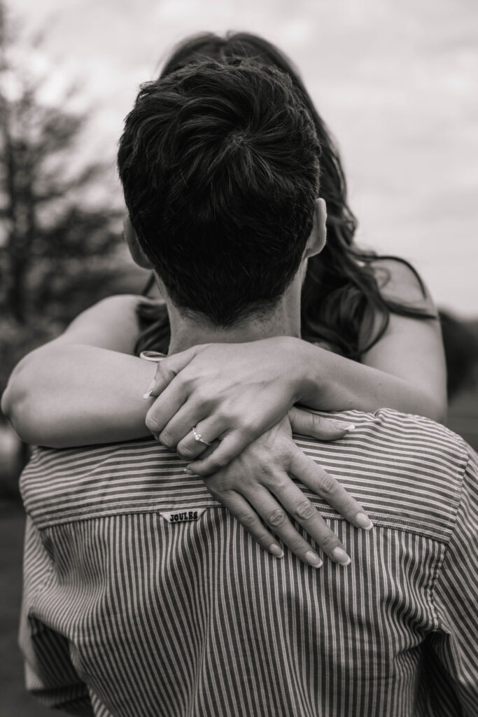 An image of a woman wearing an engagement ring hugging her partner from around his neck