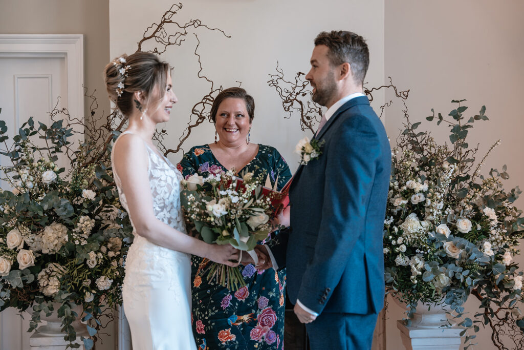 A bride and groom stand at the top of the aisle, with their celebrant smiling between them, they are surrounded by a beautiful floral arrangement
