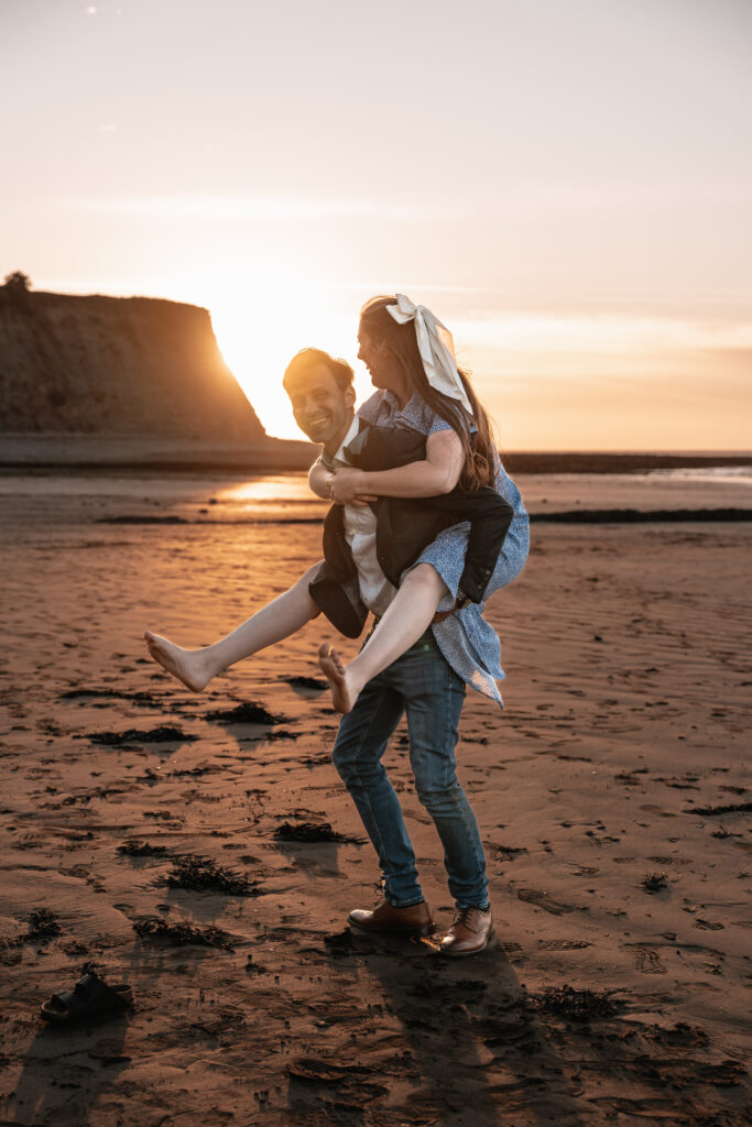 A man giving his partner a piggy back in front of the sunset on the beach