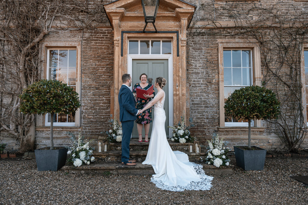 A bride and groom stand holding hands on the steps outside of their wedding venue with their celebrant smiling between 