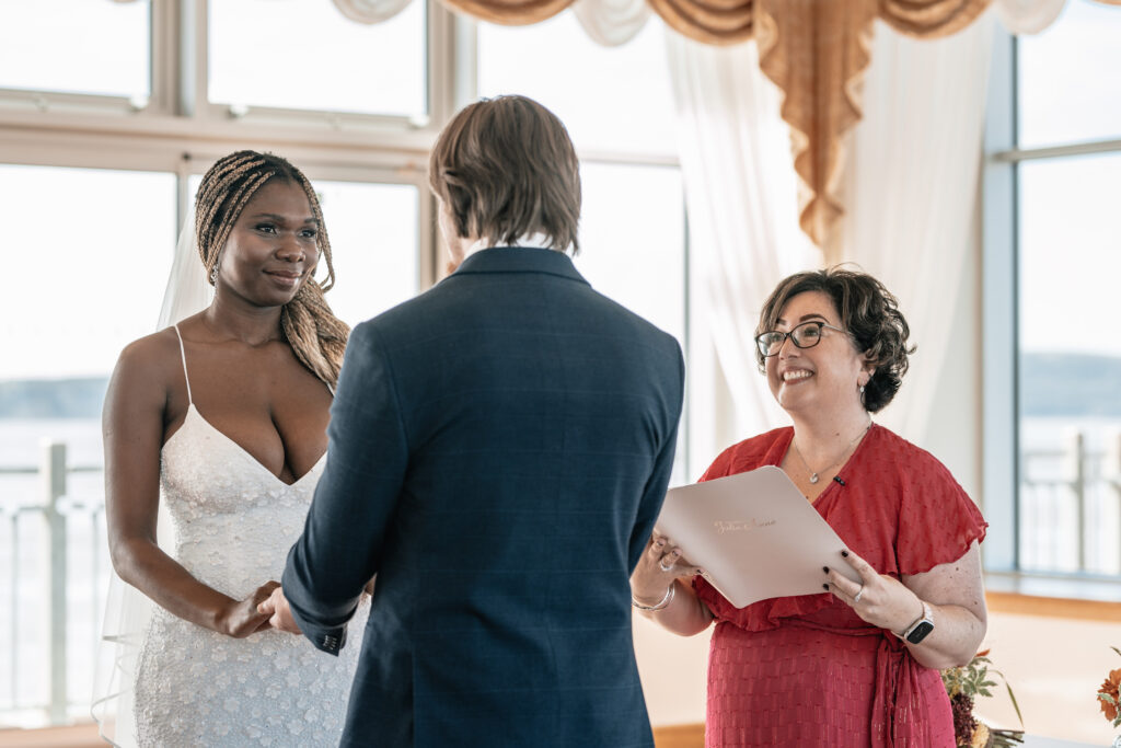 A bride looks into her groom's eyes whilst their celebrant smiles at them