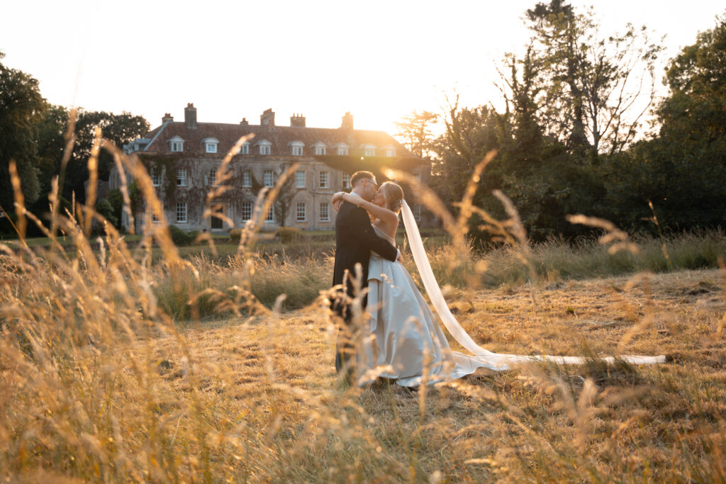 A bride and groom walk hand in hand in a sunlit field in front of Holbrook Manor. She wears a strapless gown and smiles, while he wears a black suit. 
