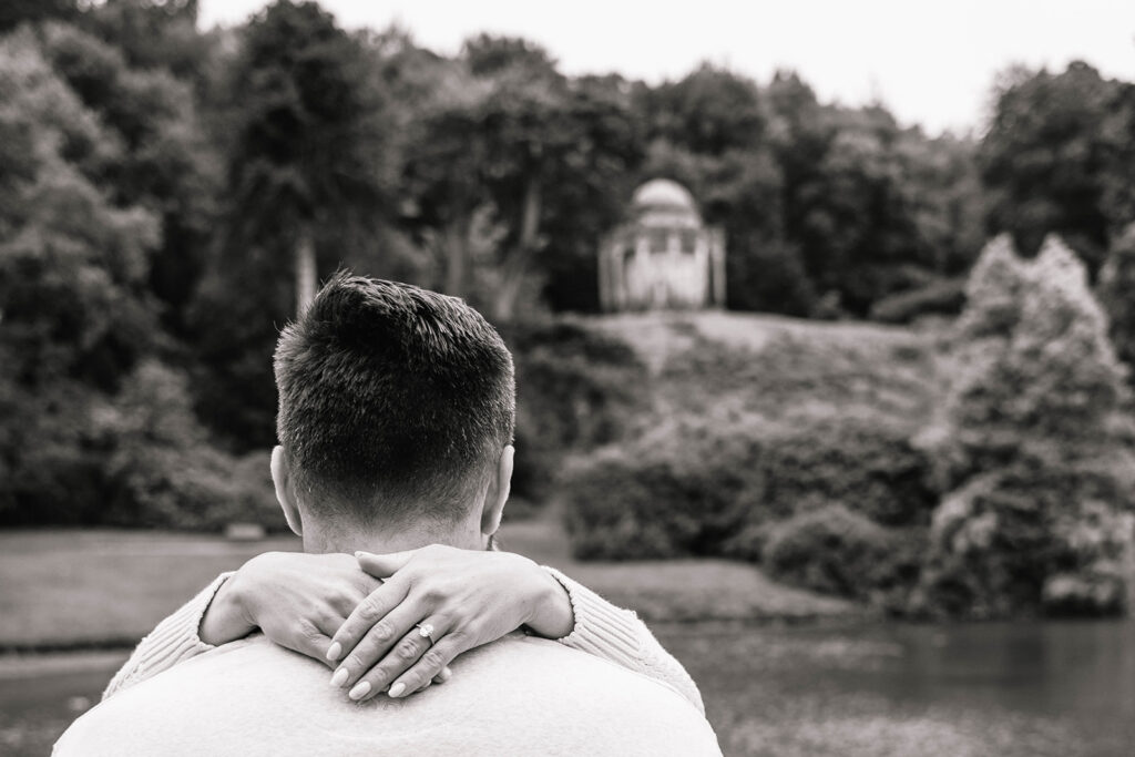 The back of a man's head with his fiance's hands around his neck showing her engagement ring. 