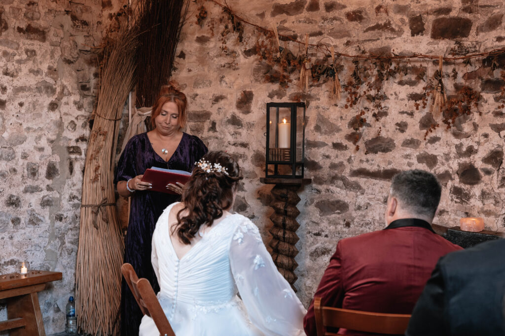 A bride and groom are seated whilst listening to their celebrant reading her script, at Hawke Barn Wedding Venue