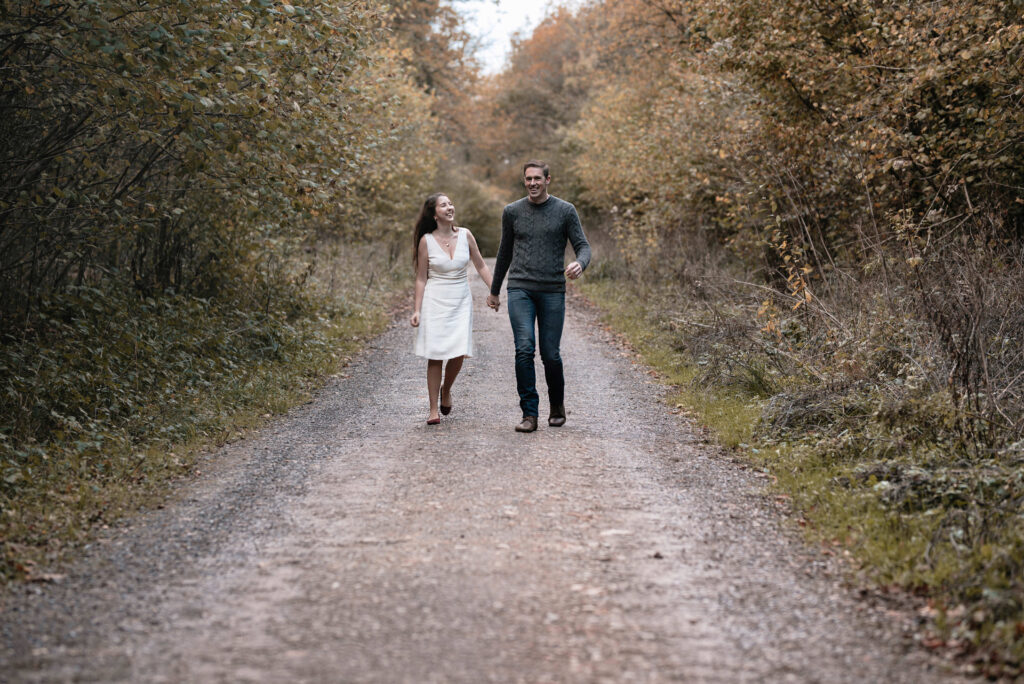 Man and woman holding hands, walking together laughing surrounded by an autumnal scene of orange trees and fallen leaves