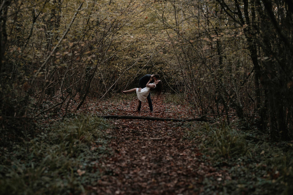 Man and woman practicing a dip dance move in a wooded area surrounded by trees and fallen leaves