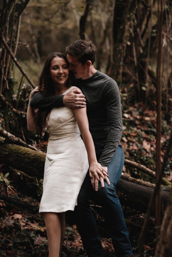 Woman sat on man’s lap on a log in wooded area, holding hands and laughing together