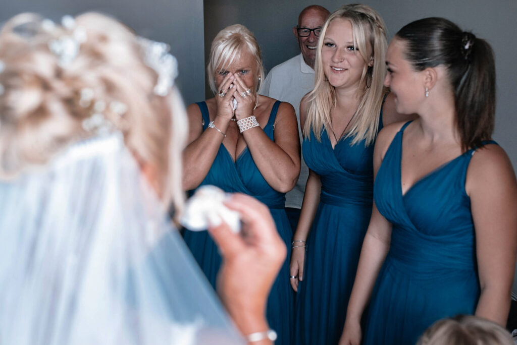 Three bridesmaids smiling and crying happy tears whilst seeing the bride for the first time. The back of the brides head is in the foreground holding a tissue to her face
