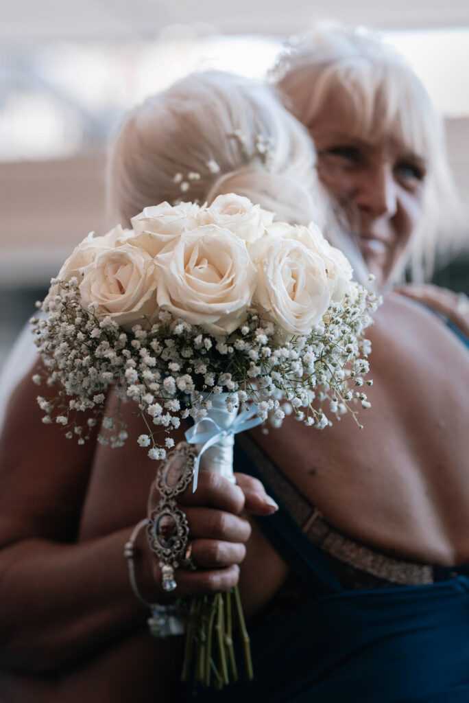 Blurred photo of bride hugging her bridesmaid. In the foreground her flowers are in focus and shows a memorial pendant with her late parents photographs