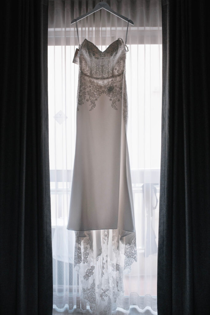 Lace detailed white wedding dress hanging from a hanger in a back-lit window framed by dark curtains 